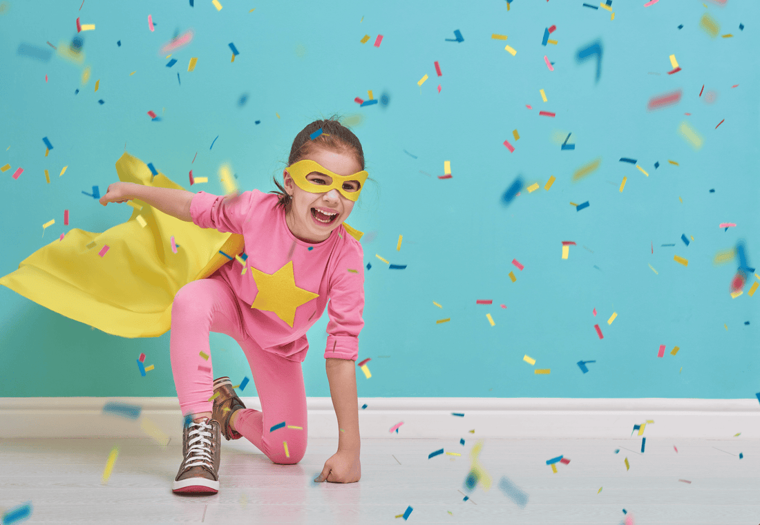 A young girl in a pink super suit with a yellow star on her chest and a yellow cape on her back. She is on one knee with a fist on the ground, a classic superhero pose. There is a baby blue background and confetti falling around her.
