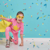 A young girl in a pink super suit with a yellow star on her chest and a yellow cape on her back. She is on one knee with a fist on the ground, a classic superhero pose. There is a baby blue background and confetti falling around her.